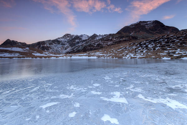 Pink lights of dawn on the snowy peaks around the frozen surface of Andossi Lake Spluga Valley Valtellina Lombardy Italy Europe