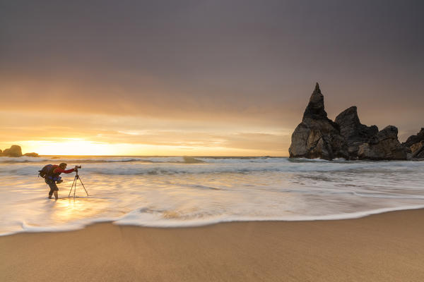 Photographer among the ocean waves under the golden sky at sunset Praia da Ursa Cabo da Roca Colares Sintra Portugal Europe