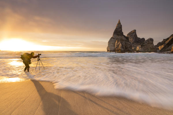 Photographer among the ocean waves under the golden sky at sunset Praia da Ursa Cabo da Roca Colares Sintra Portugal Europe