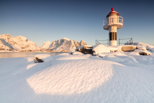 The lighthouse surrounded by snow frames the snowy peaks and the frozen sea Reine Nordland Lofoten Islands Norway Europe
