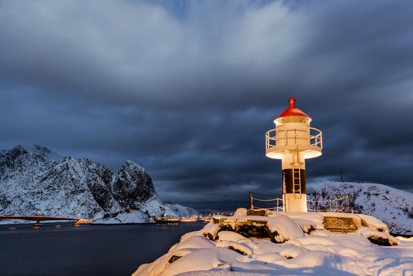 A lighthouse in the snow in the Arctic night with the village of Reine in the background Nordland Lofoten Islands Norway Europe