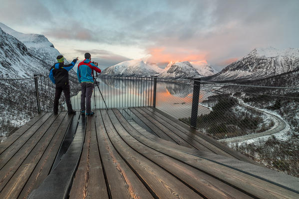 Photographers on platform admire the snowy peaks reflected in sea at sunset Bergsbotn Senja Troms County Norway Europe