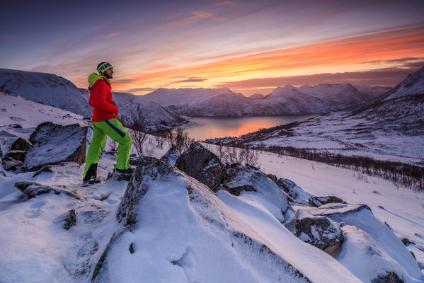 Hiker admires the frozen sea surrounded by snow framed by the orange sky at sunset Torsken Senja Troms County Norway Europe