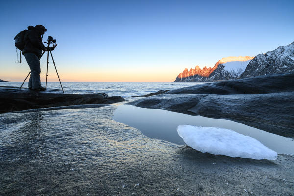Photographer on rocks admires snowy peaks and cold sea under the orange sky at dawn Tungeneset Senja Troms county Norway Europe