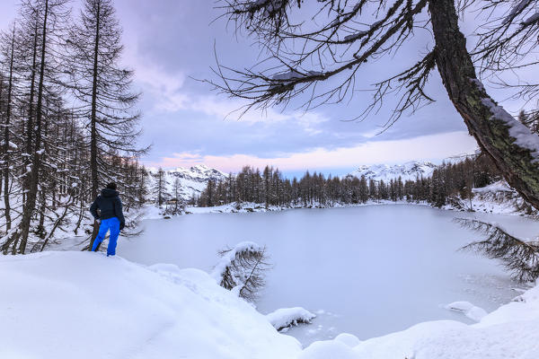 Hiker admires Lago Azzurro completely frozen at dawn Spluga Valley Province of Sondrio Valtellina Lombardy Italy Europe