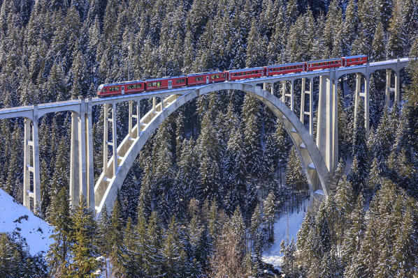 Red train of Rhaetian Railway on Langwies Viaduct surrounded by snowy woods Canton of Graubünden Switzerland Europe