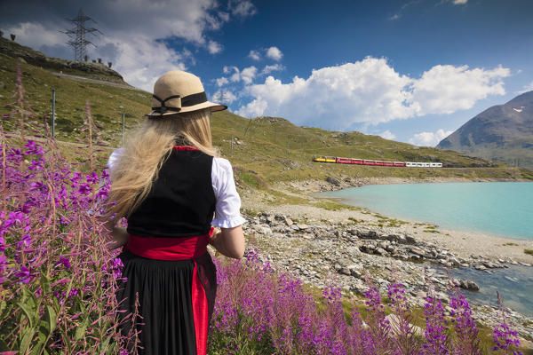 Woman with traditional clothes admires the red train at Lake Bianco Bernina Pass Canton of Graubünden Engadin Switzerland Europe