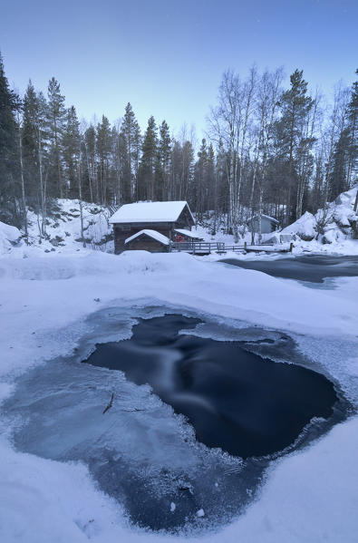 The dusk frames the frozen water in the snowy woods and the wooden hut Juuma Myllykoski Lapland region Finland Europe