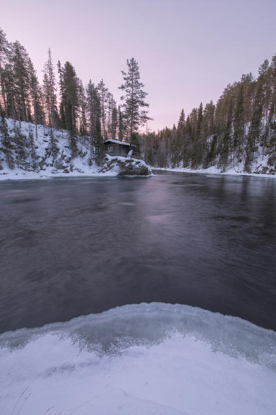 A wooden hut surrounded by the frozen river and snowy woods at dusk Juuma Myllykoski Lapland region Finland Europe