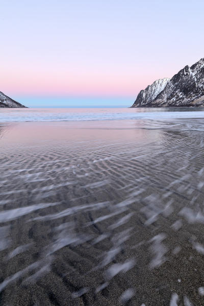 The pink light of sunrise frames the waves of frozen sea surrounded by snowy peaks Ersfjord Senja Tromsø Norway Europe