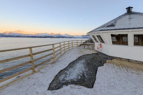 Wooden walkway and terrace on the cold sea surrounded by snowy peaks at sunset Hamn i Senja Bergsfjordens Tromsø Norway Europe