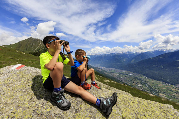 Two children sit on rocks look towards Morbegno with binoculars, Alpe Bassetta, Valtellina, Sondrio, Lombardy, Italy