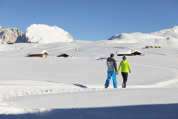 two models are walking on a winter romantic path along the Seiser Alm, with some huts and the Plattkofel Peak in the background, Bolzano province, South Tyrol, Trentino Alto Adige, Italy, Europe