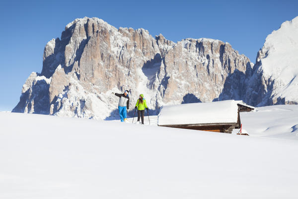 two models are walking in the fresh snow along the Seiser Alm, with some huts and the Langkofel Peak in the background, Bolzano province, South Tyrol, Trentino Alto Adige, Italy, Europe