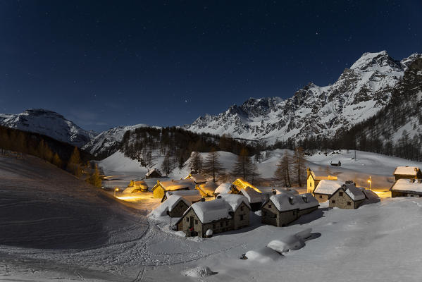 The Alp Crampiolo (Alpe Crampiolo) covered with snow in a full moon night in winter: Crampiolo is a small village in the Alpe Veglia and Alpe Devero Natural Park. The full moon light up the snowy mountains and all around the snowy village. (Baceno, Verbano Cusio Ossola province, Piedmont, Italy, Europe)