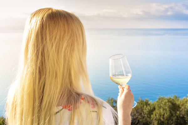 Caucasian blonde girl with a glass of wine looks the sea. Corniglia, Cinque Terre, Liguria, Italy, Europe.
