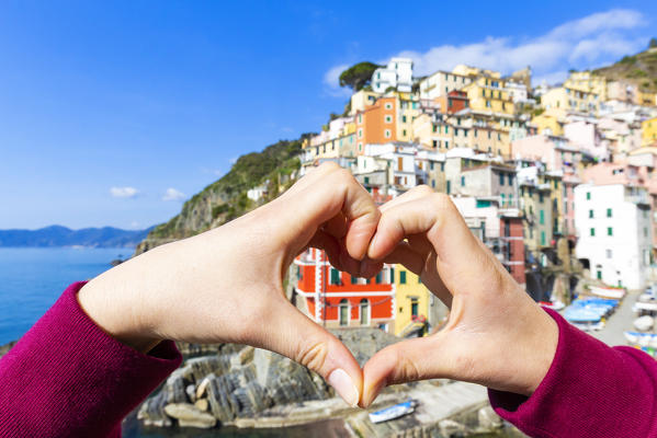 Female hand making heart shape in front the village of Riomaggiore, Cinque Terre, Liguria, Italy, Europe.