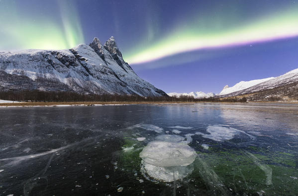 Otertinden, one of the mythical peaks of Norwegian Lapland illuminates by moon with the northern lights that colored ice bubble of a iced lake. Otertinden, Signaldalen, Storfjord, Lyngen Alps, Troms, Norway, Lapland, Europe.