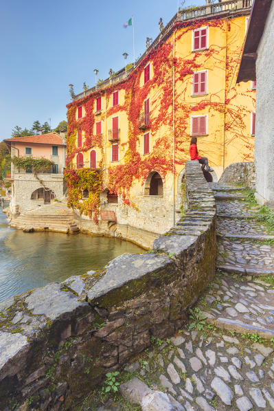 A tourist sitting on the bridge of Nesso village to admire a red ivy, Como province, Lombardy, Italy, Europe (MR)
