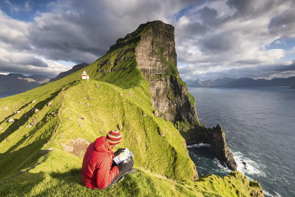 Hiker is watching the map above the ocean towards Kallur lighthouse, Kalsoy island, Faroe Islands, Denmark