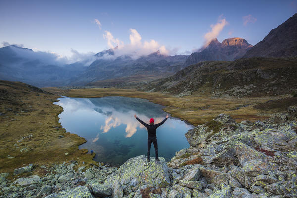 Hiker standing on lake shore with outstretched arms, Pian del Lago, Val Grosina, Valtellina, Sondrio province, Lombardy, Italy