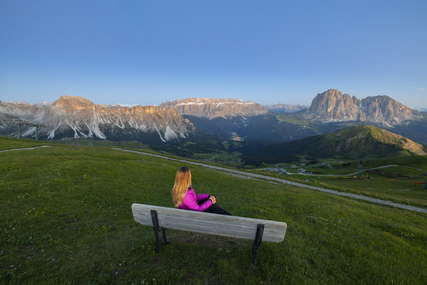 A woman sits on a bench at Seceda during dusk and observes mountains, Gruppo delle Odle, Dolomiti di Gardena, Bolzano, Trentino Alto Adige, Italy, Southern Europe 