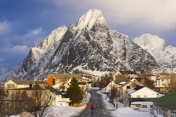A tourist stands in the main road of Reine in front of Olstinden mountain at sunset during winter, Moskenes, Moskenesoya, Nordland, Lofoten, Norway, Northern Europe 