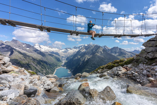 young woman sitting on the suspension bridge near the olperer hut with Schlegeispeicher lake on the background, Zillertal Alps, Tyrol, Schwaz district, Austria.